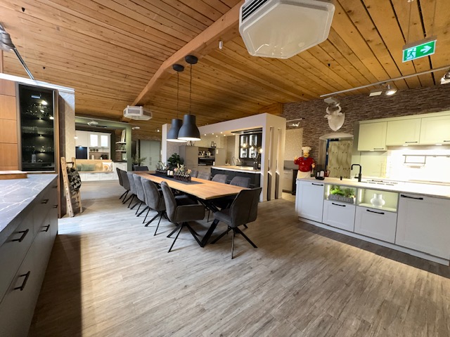 Moderne Küche und Essbereich mit einem langen Holztisch, schwarzen Stühlen, Pendelleuchten, einer holzvertäfelten Decke und weißen Schränken.
